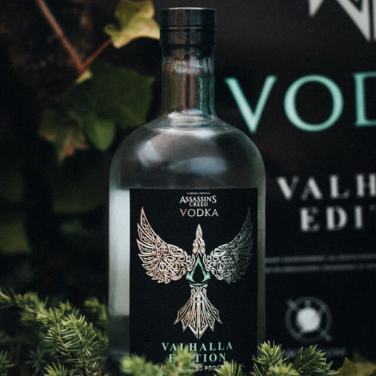 A bottle of Assassin's Creed Vodka Valhalla Edition, featuring a label with a metallic angel or bird design, set against a natural background with foliage.